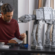 A white man in a burgendy shirt looks down at some Lego pieces as a Star Wars AT-AT Lego creation stands tall on a table in front of him.